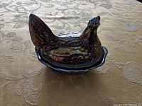 Side view of the Fenton carnival glass hen covered dish on a patterned beige tablecloth showing the iridescent finish and form.
