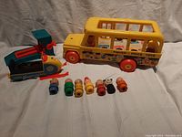 Yellow Fisher Price school bus with red wheels and open windows, with some wooden Little People figures inside seen through windows alongside several Little People figures lined up in front of the bus.