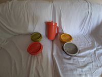 Full view of vintage Tupperware orange jug and four circular containers with lids in various colors on white fabric background.
