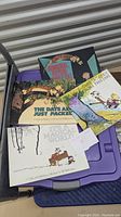 Four Calvin & Hobbes comic books shown fanned out on a purple storage bin. Visible titles include Homicidal Psycho Jungle Cat, The Days Are Just Packed, Sunday Pages 1985-1995, and It's a Magical World. The books have illustrated cartoon covers with colorful artwork featuring Calvin and Hobbes characters.