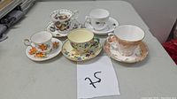 Five assorted tea cups with matching saucers arranged in a row showing tops and sides, placed on a gray table with lot number 75.