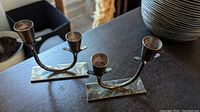 Photo of two silver-toned metal candleholders with square bases on a dark surface, showing tarnish and curved arms designed to hold two candles each