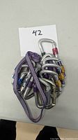 Group of eight assorted locking metal carabiners with colored locking mechanisms and one purple patterned climbing sling laid on table