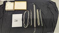 Photograph showing all assorted jewelry pieces laid on a black fabric background with a numbered card showing '26'. Included are a multi-strand pearl necklace, rhinestone bracelet, pearl bracelet, linked silver-tone bracelet, and an open empty jewelry box.
