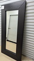 Framed large rectangular mirror standing upright against a corrugated metal wall, showing the dark wood frame with mitered corners.