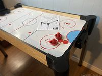 Full view of air hockey table showing wood frame, white playing surface with rink markings, black corner protectors, metal legs, red pushers, and puck on the surface, manual present.