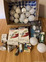 Box containing a variety of white and frosted round bulbs and some bulbs in original Philips and other brand packaging. Also includes packaged fuses/mini-breakers and a variety of loose bulbs.