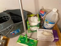 Swiffer mop, mop bucket, disposable dry wipes boxes, vinyl gloves box, partially full Zep glass cleaner placing overall lot contents.