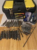 Black and yellow Stanley toolbox with a variety of drill bits spread on the wooden surface in front of it. The drill bits vary in size and type.