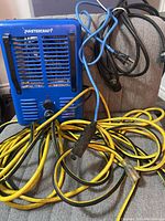 Blue Mastercraft heater with ventilation grill and control knob, along with tangled yellow, blue, and black extension cords on a couch.