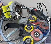 Photo showing Ryobi circular saw with yellow handle, Jobmate angle grinder, and multiple grinding discs spread on a clear glass table.