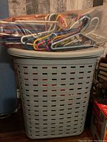 Grey plastic laundry hamper with a lid and a large bundle of colorful kids plastic hangers on top, placed next to a wooden surface and other household items.