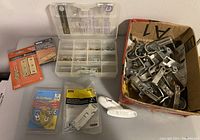 Overview image showing assorted hinges, curtain rod brackets in a box, wire hangers, hooks, and a plastic storage container on a table.