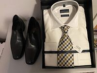 Pair of black leather men's shoes beside white men's dress shirt with gold and blue plaid tie, all boxed.