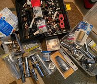 Assorted plumbing components scattered on carpet and inside plastic containers, including chrome drain assemblies, brass valves, small fittings, and a toolbox with numerous small items.