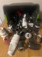 Large open container with assorted plumbing parts including multiple black PVC fittings, white and red couplings and valves, and metal hose clamps spread on table.