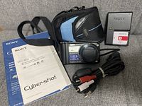 Photo shows the Sony Cyber-shot DSC-H55 digital camera with a sticker on lens indicating features; Roots branded carrying case; instruction manual and secondary booklet; a USB cable and RCA AV cable coiled together.