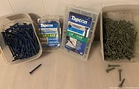 Plastic containers and loose screws of blue coated Tapcon screws and gray coated masonry screws arranged on a flat surface.