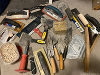 Photo shows an assortment of masonry and tiling hand tools including trowels, chisel, floats, and an anvil. A container of loose tile spacers is visible.