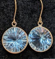 Close-up front view of pair of blue topaz drop earrings with 10K gold hooks and settings shown against black background.