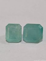 Close-up front-facing image of two square or rectangular step-cut emerald stones side by side on a plain white background.