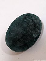 Close-up photo of a dark green oval emerald showing natural inclusions and veining on a white background.