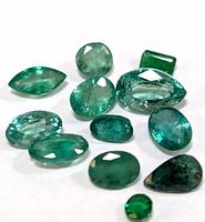 Photo of 11 loose genuine emerald gemstones on a white background showing their sizes, cuts, and coloration.