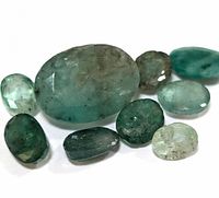 Photo showing nine various shaped and sized emerald gemstones with green hues and natural inclusions on a white background.