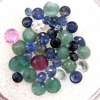 Close-up photo showing assorted loose gemstones including green emeralds, blue sapphires, a pink stone, a deep red ruby, and colorless faceted stones on a white background.