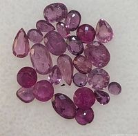 Close-up front view of multiple loose faceted ruby gemstones scattered on a flat surface showing different shapes and pink to purple-red hues.