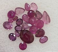 Collection of 23 assorted cut ruby gemstones displayed on white background, showing variation in cuts and color depths.