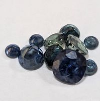 Multiple faceted sapphire gemstones lying on a white background, showing a variety of shapes including round, cushion and marquise cuts, in blue and green hues.