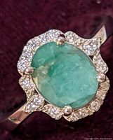 Close-up view of oval emerald stone with clear cubic zirconia halo set in sterling silver ring on dark velvet background.