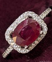 Close-up image of sterling silver ring with oval ruby center stone surrounded by cubic zirconia halo and cubic zirconia stones on the band.
