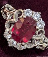 Close-up front view showing oval ruby center and clear stone halo with scrollwork sterling silver band.