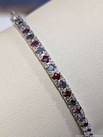Close-up angle of the colorful ruby and sapphire stones embedded in a thin sterling silver bangle bracelet resting on a white cushion.