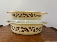 Two Pyrex Golden Acorn pattern casserole dishes stacked, showing the brown leaf and acorn vine design on cream background with clear glass lids.