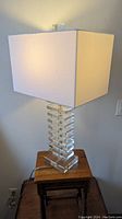 Crystal glass table lamp with stacked rectangular block base and white rectangular shade on wooden stool, lit to show illumination