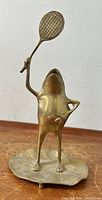 Brass frog figurine holding tennis racket, standing on lily pad, front view