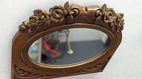 Front view showing the carved wood floral top and oval mirror glass reflecting an indoor scene.