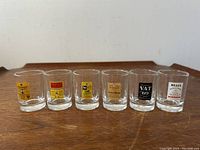 Front view of six vintage clear glass shot glasses each with unique colorful enameled Scotch whiskey advertising labels arranged in a row on a wooden surface.