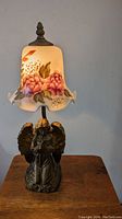 Front view of vintage figural guardian angel table lamp with reverse painted floral art glass shade, showing the lamp illuminated and details of the angel base and painted glass shade.