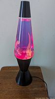 Full view of the lava lamp turned on, showing pink wax blobs moving in purple liquid inside a conical clear glass container. Black base and cap visible.