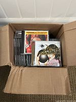 Open cardboard box showing multiple CD cases standing upright including 'Commodores All The Great Hits' and 'Hoobastank' albums on top.