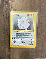 Front view of a holographic Chansey Pokémon card showing details such as HP, moves, and stats on a wooden surface.