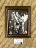 Framed 8x10 black and white photo of Eddie Shack holding Stanley Cup, signed by Eddie Shack with visible signature and plaques inside frame, displayed on carpet.
