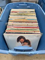 Photo showing blue plastic container filled with about 40 vintage vinyl records. Album covers show multiple different colors and design styles. Top record cover depicts a man and woman embracing in a warm vintage portrait photograph.