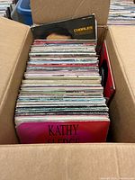 Open cardboard box filled with approximately 50 vintage vinyl LP records. Front record titled 'Kathy Sledge' visible, another titled 'Charles Aznavour' near the back.