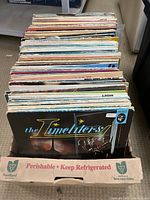 Photo showing a collection of approximately 30 vintage vinyl records in their sleeves standing upright in a cardboard box. The front visible record is titled 'The Timeliters'.
