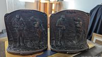 Pair of solid cast iron bookends titled 'The Gleaners' showing front embossed figural scene with two gleaners in rural setting, mounted vertically.